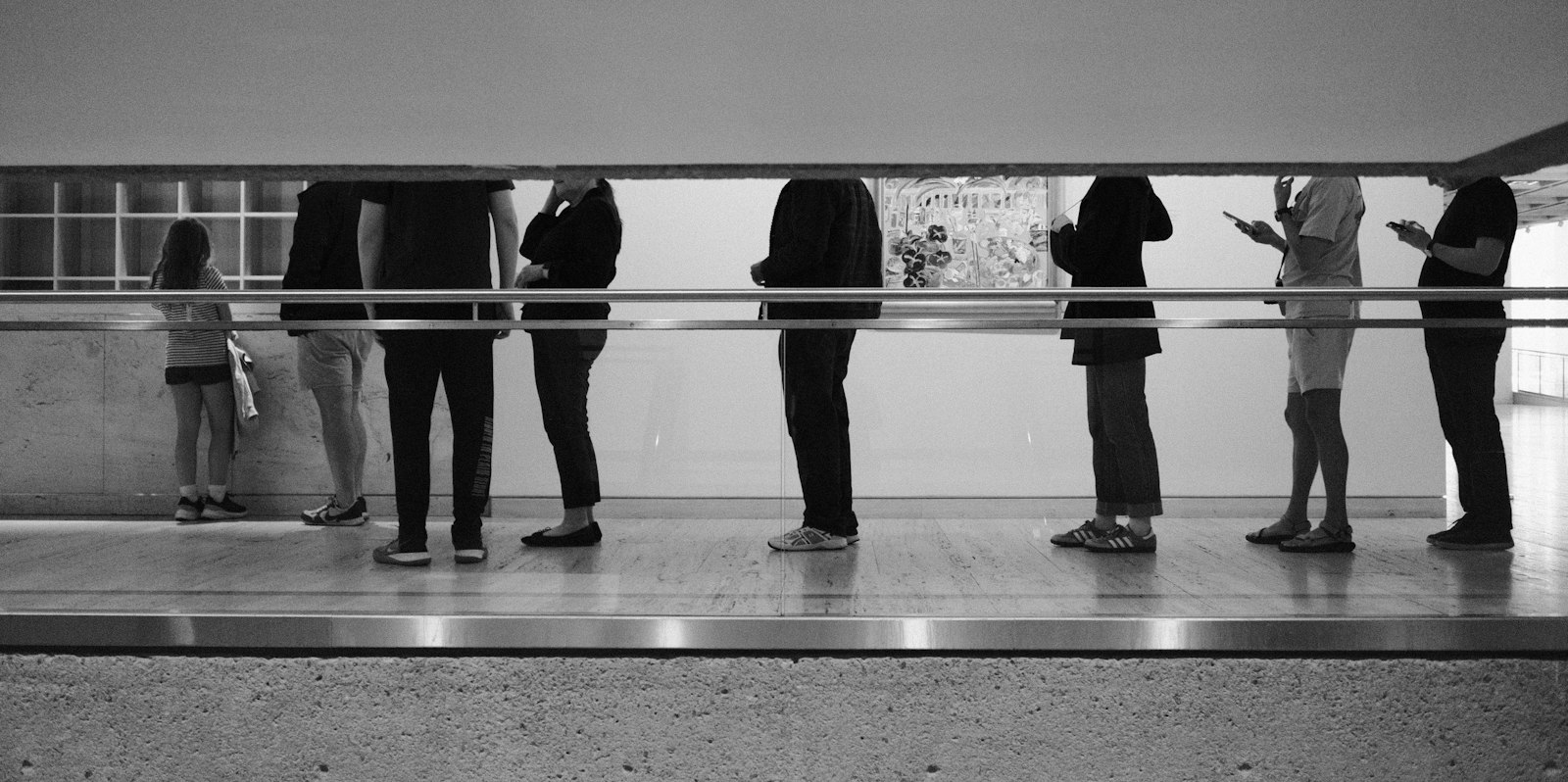 Una fila de personas en un espacio interior. El encuadre deja fuera sus caras: se ven cuerpos, bolsos y celulares.